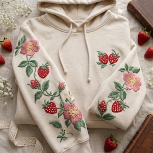 May include: A cream-coloured hooded sweatshirt featuring intricate embroidery of pink flowers, green leaves, and red strawberries on both sleeves. A small matching strawberry embroidery is positioned on the upper right chest area of the garment.