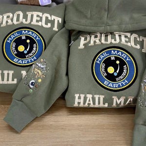 May include: Two olive green hooded sweatshirts featuring the text PROJECT HAIL MARY embroidered in cream. A circular patch displays the text HAIL MARY GRACE EARTH with space-themed graphics. The sleeves feature embroidered space motifs.