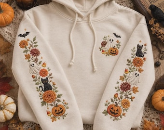 May include: A cream-colored hooded sweatshirt featuring intricate embroidery on both sleeves and the chest. The designs include orange pumpkins, white ghosts, black cats, autumn flowers, leaves, bats, and candy corn in vibrant seasonal colors.