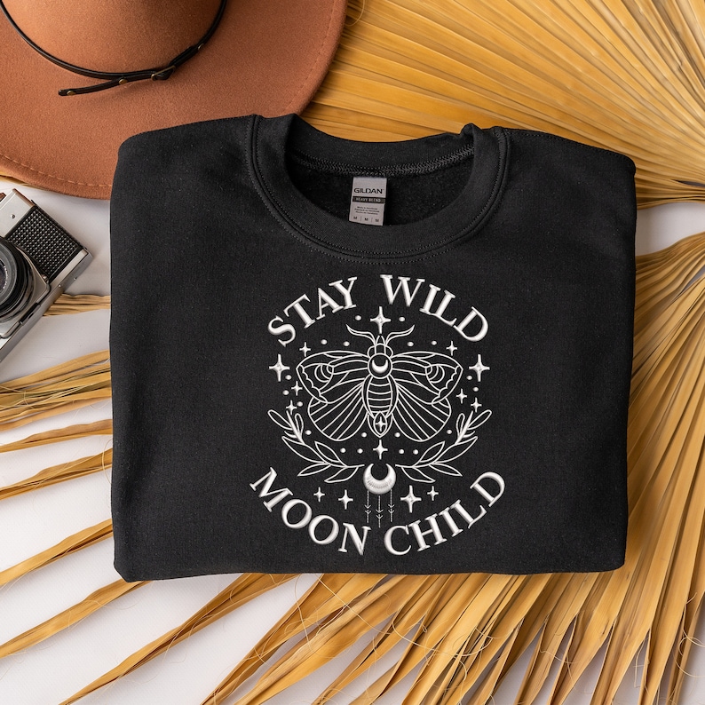 Puede incluir: Sudadera negra con texto blanco que dice "Stay Wild Moon Child" y un gr&aacute;fico de una polilla con una luna creciente y estrellas.