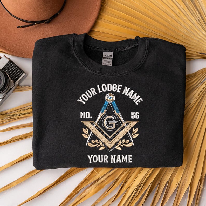 May include: Black crewneck sweatshirt with a Masonic emblem design. The emblem features a compass, square, and the letter "G" in the center, with text that reads "Your Lodge Name", "No. 56", and "Your Name".