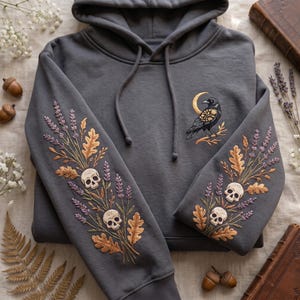 May include: A charcoal gray hooded sweatshirt featuring intricate embroidery. The sleeves display a botanical design of lavender sprigs, oak leaves, and two skulls. The chest area features a black crow perched on a branch with a gold crescent moon.