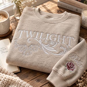 May include: A beige crewneck sweatshirt featuring white embroidered text that reads TWILIGHT across the chest with decorative scrollwork underneath. The sleeve includes the embroidered name Bella above a detailed red and green rose graphic.