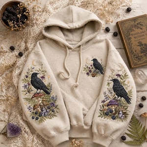 Könnte beinhalten: Ein beigefarbener Kapuzenpullover mit detaillierten Stickereien an den Ärmeln und auf der Brust. Das Design zeigt schwarze Raben auf Pilzen, umgeben von violetten Blumen, grünen Farnen, Brombeeren, Kristallen und Mondsichelmotiven.