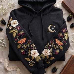 Puede incluir: Una sudadera negra con capucha que presenta un bordado intrincado en ambas mangas y en el pecho. Las mangas muestran flores blancas, hojas verdes, bayas oscuras y polillas naranjas. El pecho presenta un diseño de luna creciente y estrella.