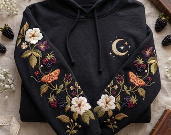 May include: A black hooded sweatshirt featuring intricate embroidery on both sleeves and the chest. The sleeves display white flowers, green leaves, dark berries, and orange moths. The chest features a crescent moon and star design.