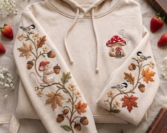 May include: A cream-colored hooded sweatshirt featuring intricate embroidery on the sleeves and chest. The sleeves display a detailed scene of a mouse, acorns, autumn leaves, and birds. The chest features a small embroidered mushroom house.