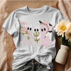 May include: White t-shirt featuring embroidered ghosts holding flowers, sprigs, and bats. The ghosts are in shades of white and pink, with black oval eyes. The design is a whimsical, Halloween-themed graphic.