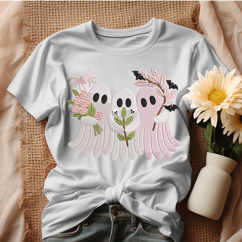 May include: White t-shirt featuring embroidered ghosts holding flowers, sprigs, and bats. The ghosts are in shades of white and pink, with black oval eyes. The design is a whimsical, Halloween-themed graphic.
