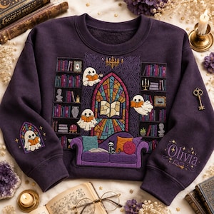 May include: A dark purple crewneck sweatshirt featuring an embroidered library scene with ghosts reading books, a stained glass window, a purple sofa, and a black cat. The left sleeve displays a ghost reading, and the right sleeve features the name Olivia.