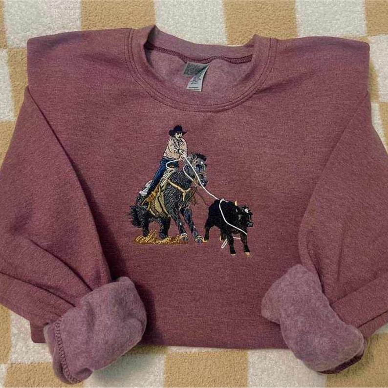 Pode incluir: Uma camisola de cor castanho-avermelhada com um desenho bordado de uma pessoa a montar a cavalo e a la&ccedil;ar um bezerro. O desenho &eacute; em tons de castanho, preto e branco.