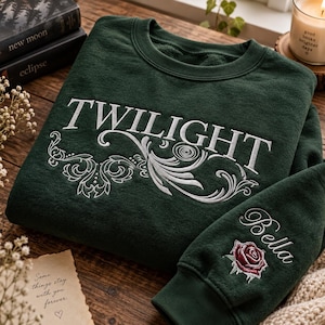 May include: A dark forest green crewneck sweatshirt featuring white embroidered text that reads TWILIGHT above a decorative white floral scroll design. The sleeve features the embroidered name Bella in white script above a red rose graphic.