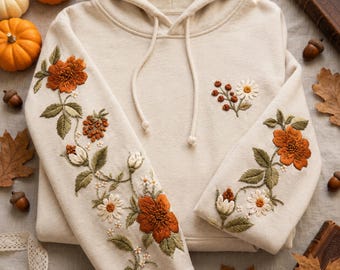 May include: A cream-colored hooded sweatshirt featuring intricate embroidery of orange flowers, green leaves, and small white blossoms along the sleeves and a small floral cluster on the chest. The hoodie includes matching cream-colored drawstrings.