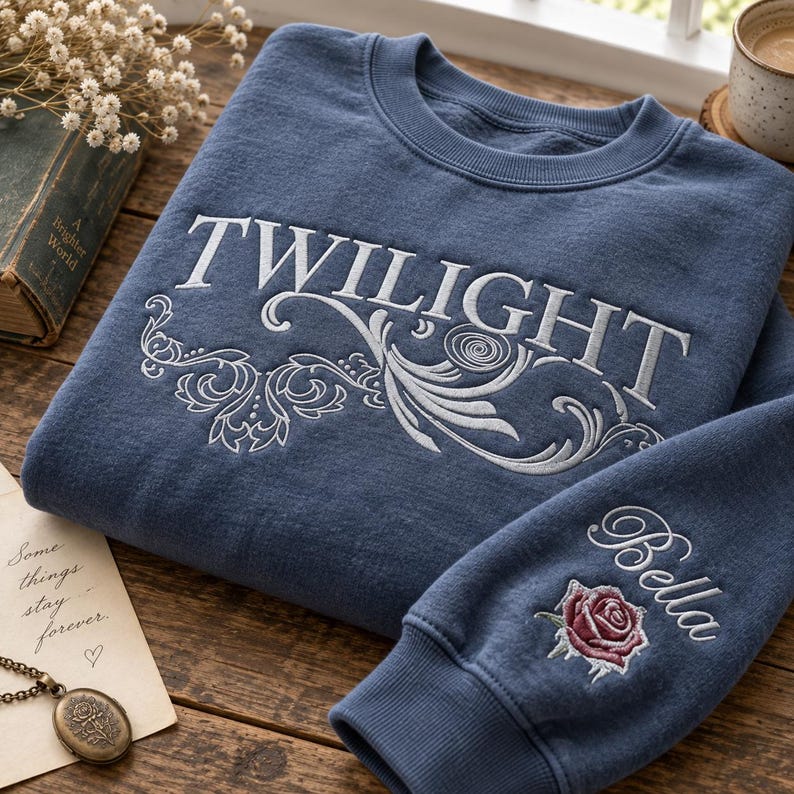 May include: A blue crewneck sweatshirt featuring the word TWILIGHT embroidered in white across the chest with white decorative scrollwork underneath. The sleeve features the name Bella embroidered in white above a red and green rose graphic.