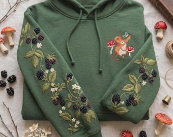 May include: A forest green hooded sweatshirt featuring detailed embroidery of blackberry vines with dark purple berries and white flowers on both sleeves. The chest displays a small embroidered mouse standing between two red and white mushrooms.