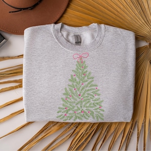 May include: A light gray sweatshirt featuring a green embroidered Christmas tree design with pink ornaments and a pink bow at the top. The sweatshirt is folded and displayed on a neutral background.
