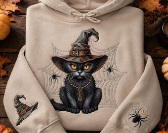 May include: A beige hooded sweatshirt featuring a graphic of a black cat wearing a witch hat, centered in a spider web. The cat wears a gold chain necklace. The left sleeve displays a witch hat, and the right sleeve shows a spider on a web.