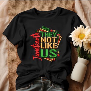 May include: Black t-shirt with a graphic design that says "Juneteenth 1865 They Not Like Us" in colorful letters with stars.