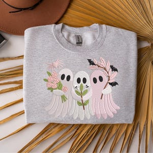May include: A light gray sweatshirt featuring an embroidered design of three ghost figures. The ghosts are in shades of pink and white, holding flowers, leaves, and bats. The sweatshirt is folded on a neutral surface.