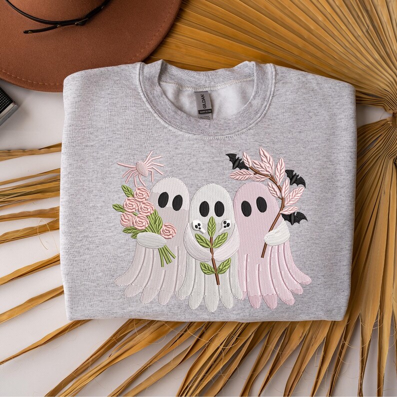 May include: A light gray sweatshirt featuring an embroidered design of three ghost figures. The ghosts are in shades of pink and white, holding flowers, leaves, and bats. The sweatshirt is folded on a neutral surface.