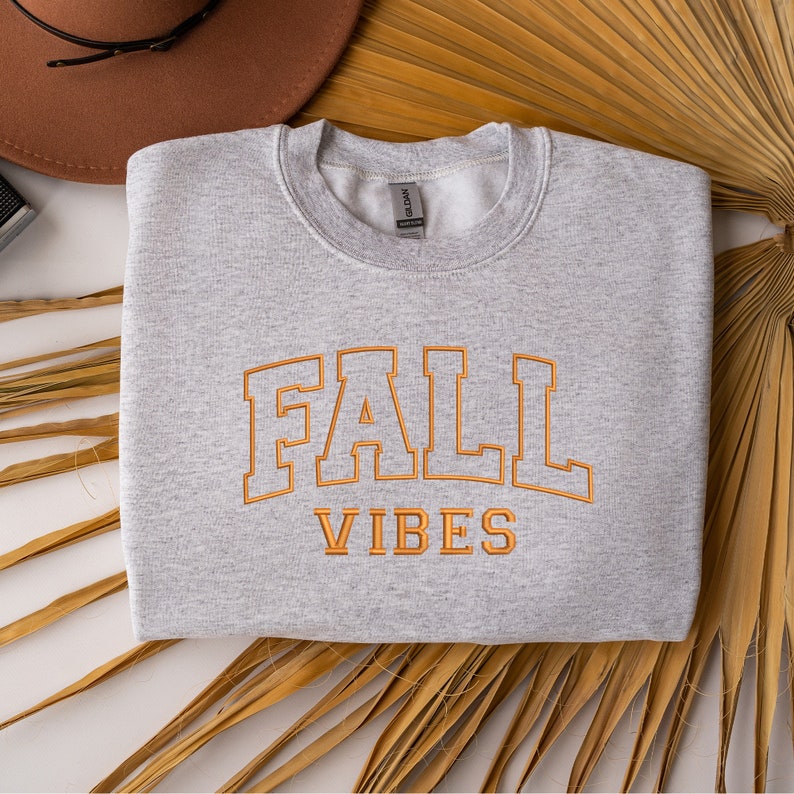 May include: A light gray sweatshirt with the words "FALL VIBES" embroidered in orange. The sweatshirt is folded and placed on a white surface with a brown hat and dried palm leaves in the background.