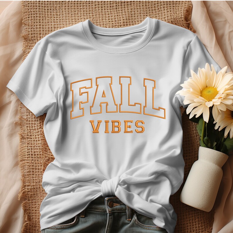 May include: White t-shirt with the words "FALL VIBES" in orange block letters. The shirt is knotted at the waist. A vase of yellow flowers sits to the right of the shirt.