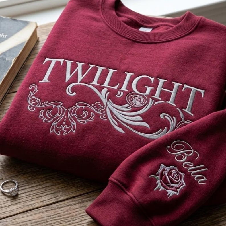May include: A maroon crewneck sweatshirt featuring the word TWILIGHT embroidered in white capital letters across the chest, accented by white floral scrollwork. The sleeve displays the name Bella in cursive script above a white embroidered rose.
