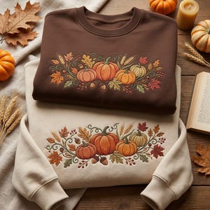 May include: A dark brown crewneck sweatshirt and a cream-coloured crewneck sweatshirt, each featuring an embroidered design of orange pumpkins, wheat stalks, acorns, and autumn leaves in shades of red, orange, yellow, and green across the chest.