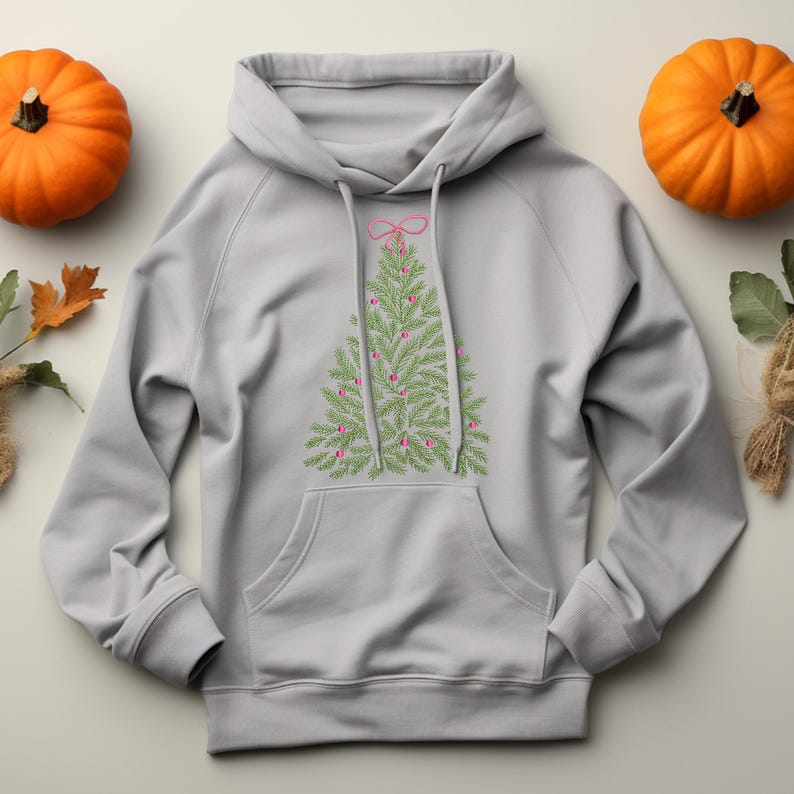 May include: A light gray hoodie featuring a green Christmas tree design with pink ornaments and a pink bow at the top. The hoodie has a kangaroo pocket and a drawstring hood. Two orange pumpkins are on either side of the hoodie.