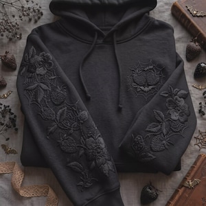 Puede incluir: Una sudadera con capucha negra que presenta bordados negros tonales en las mangas y el pecho. El bordado representa intrincados patrones florales, motivos de fresas y telarañas. La sudadera incluye cordones negros a juego en el cuello.
