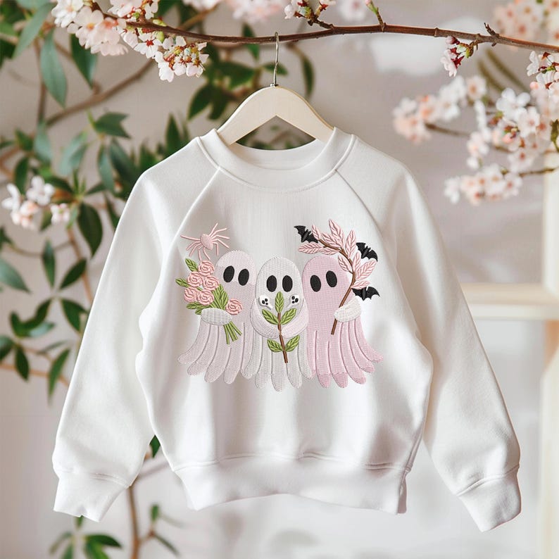May include: White sweatshirt with a crew neck and long sleeves. The front features an embroidered design of three ghost figures in white and pink, each holding flowers or branches. The design includes bats and floral accents, perfect for Halloween or spooky season.