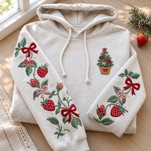 May include: A light heather gray hooded sweatshirt featuring intricate embroidery on both sleeves and the chest. The sleeves display red strawberries, green leaves, and red bows. The chest features a small embroidered Christmas tree in a basket.