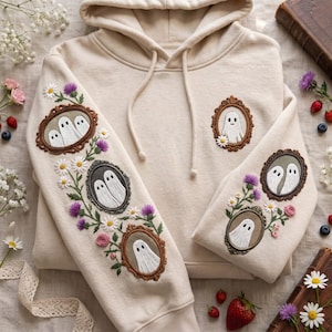Könnte beinhalten: Ein beigefarbener Kapuzenpullover mit aufwendigen Stickereien von geisterhaften Motiven in ovalen Rahmen und floralen Mustern. Die Ärmel und die Brust zeigen detaillierte braune Rahmen mit weißen Geisterfiguren, akzentuiert durch gestickte Gänseblümchen, violette Disteln und rosa Rosen.