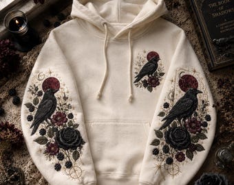 Peut inclure: Un sweat à capuche blanc cassé avec des broderies détaillées sur les manches et la poitrine gauche. Le motif représente des corbeaux noirs, des lunes rouge foncé, des roses violettes et des mûres, agrémenté de motifs d'étoiles dorées.