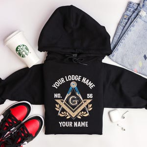 May include: Black hooded sweatshirt with a Masonic emblem design. The design includes a compass, square, and the text "YOUR LODGE NAME", "NO. 56", and "YOUR NAME". The sweatshirt is displayed with a coffee cup, sneakers, and jeans.