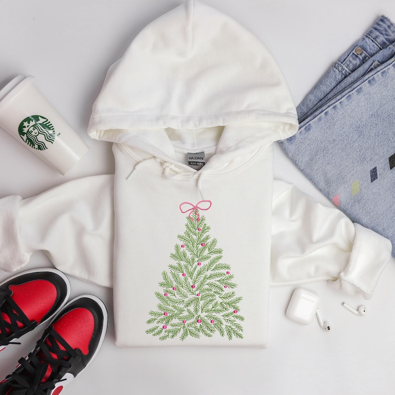 May include: A white hooded sweatshirt featuring a green Christmas tree design with pink ornaments and a pink bow at the top. The sweatshirt is on a white surface with a Starbucks cup, sneakers, and jeans.