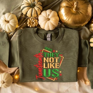 May include: A green sweatshirt with the text "Juneteenth 1865 They Not Like Us" in red, gold, and green lettering. The sweatshirt is surrounded by white and gold pumpkins.