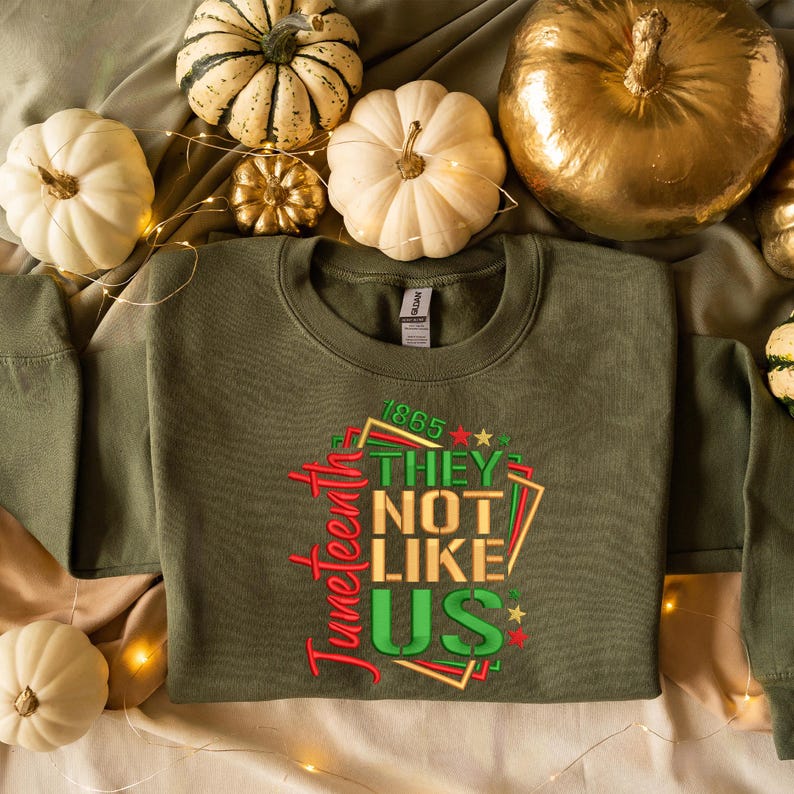 May include: A green sweatshirt with the text "Juneteenth 1865 They Not Like Us" in red, gold, and green lettering. The sweatshirt is surrounded by white and gold pumpkins.