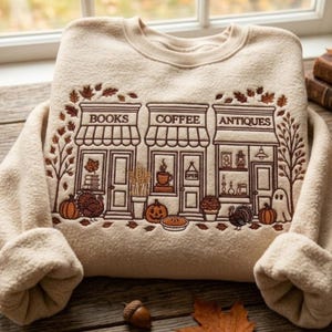 May include: A beige crewneck sweatshirt featuring brown embroidery of three storefronts labelled BOOKS, COFFEE, and ANTIQUES. The design includes autumn motifs like pumpkins, turkeys, falling leaves, a ghost, and a pie, creating a seasonal scene.