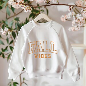 May include: White sweatshirt with the words "FALL VIBES" in orange block letters. The sweatshirt is hanging on a wooden hanger. The background has a floral arrangement.