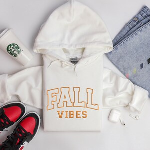 May include: A cream-colored hoodie with the words "FALL VIBES" in orange block letters. The hoodie is laid flat, with a pair of red and black sneakers, a pair of blue jeans, and a Starbucks cup nearby. The hoodie has a drawstring hood.