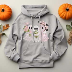 May include: A light gray hoodie with a front pocket and drawstring. The hoodie features an embroidered design of three cartoon ghosts holding flowers, leaves, and a bat. Two orange pumpkins are on either side of the hoodie.