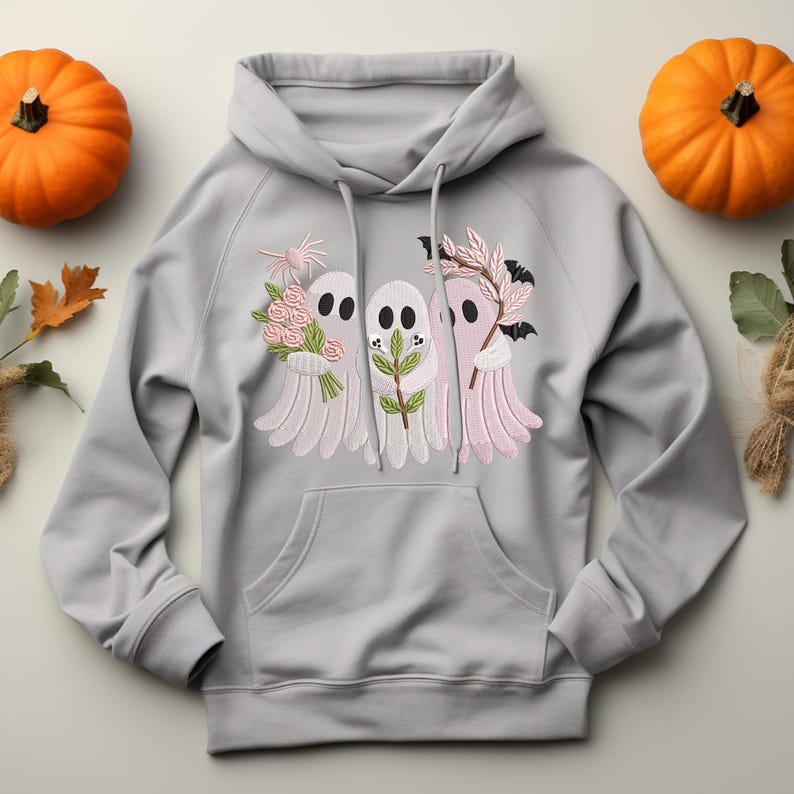 May include: A light gray hoodie with a front pocket and drawstring. The hoodie features an embroidered design of three cartoon ghosts holding flowers, leaves, and a bat. Two orange pumpkins are on either side of the hoodie.