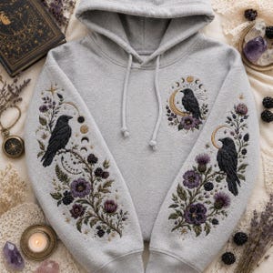 May include: A light gray hooded sweatshirt featuring detailed embroidery of black crows perched on branches with purple flowers, blackberries, and golden moon phases. The design is mirrored on both sleeves and includes a small crow motif on the chest.
