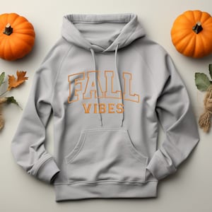 May include: A light gray hoodie with the words "FALL VIBES" in orange lettering. The hoodie has a front pocket and a drawstring hood. Two orange pumpkins and fall foliage are arranged around the hoodie.
