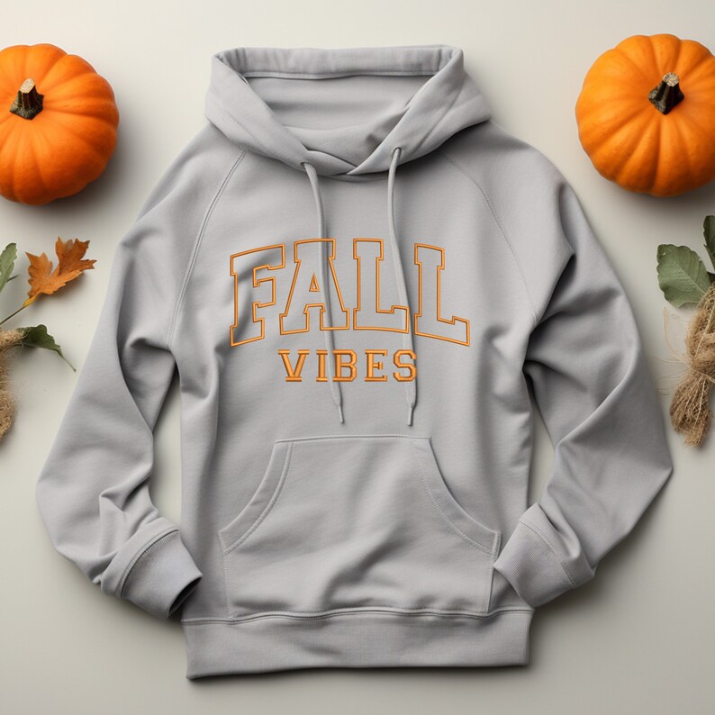May include: A light gray hoodie with the words "FALL VIBES" in orange lettering. The hoodie has a front pocket and a drawstring hood. Two orange pumpkins and fall foliage are arranged around the hoodie.