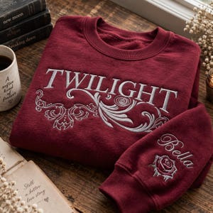 May include: A deep maroon crewneck sweatshirt featuring white embroidered text that reads TWILIGHT across the chest with decorative swirling filigree patterns. The sleeve includes the embroidered name Bella above a detailed red rose graphic.
