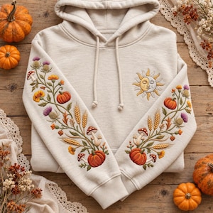 Peut inclure: Un sweat à capuche beige avec une broderie complexe sur les manches et la poitrine. Les manches présentent un motif botanique avec des citrouilles, des champignons, du blé et des chardons violets. La poitrine arbore un emblème soleil et lune.