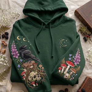 May include: A dark forest green hooded sweatshirt featuring intricate embroidery on the sleeves. The right sleeve displays a black crow perched on a tree stump with a skull, mushrooms, and purple flowers. The left sleeve features mushrooms, purple flowers, and a small skull. A gold crescent moon is embroidered on the chest.