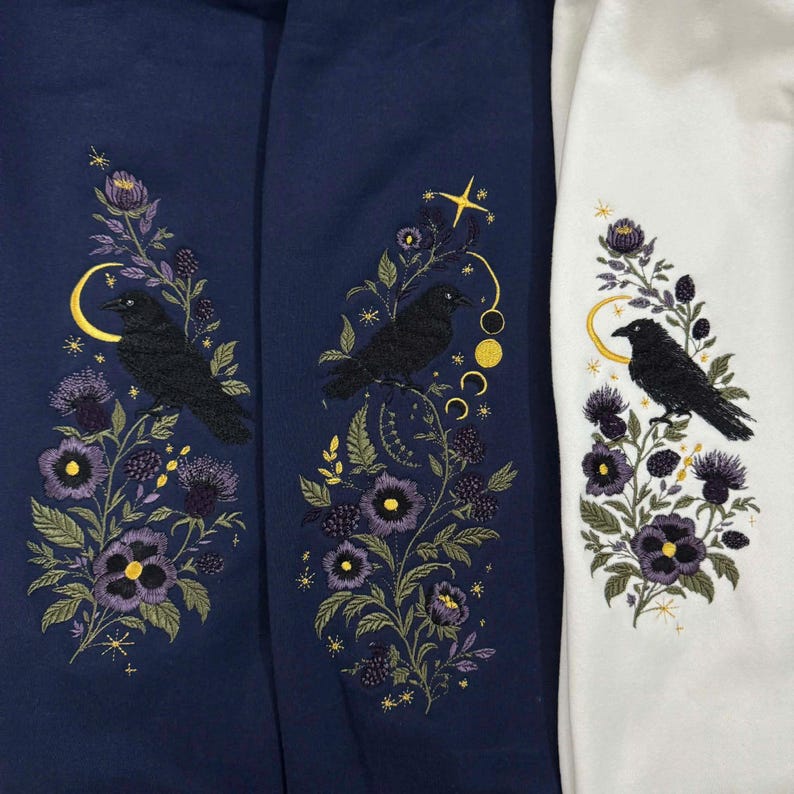 May include: Three sweatshirts, two navy blue and one white, each featuring an embroidered design of a black crow perched on a floral stem with purple flowers, green leaves, and yellow celestial elements including moons, stars, and planetary phases.
